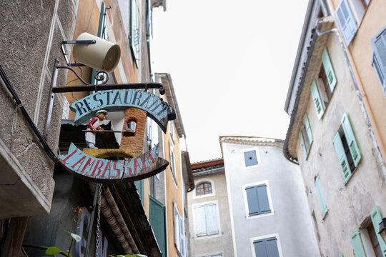 Entrevaux, France - December 26, 2025: A rustic restaurant sign with a chef figure hangs over a narrow historic street.