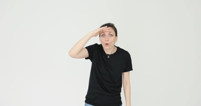 Woman in simple black T-shirt places hand against forehead. Female looks into remote distance with curious expression against white background