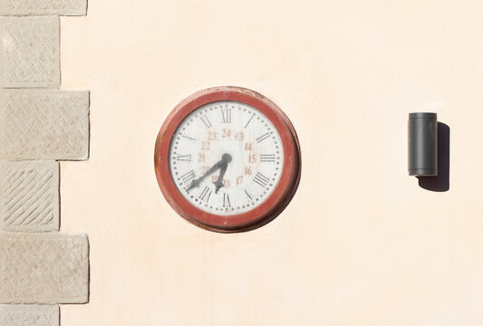 A round wall clock with Roman numerals and a small black lamp on a building wall.