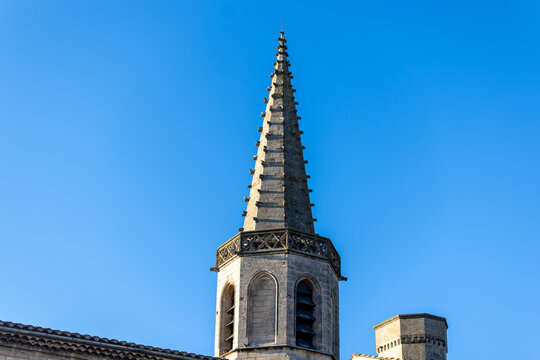 Arles, France -A tall stone bell tower with a conical spire rises against a clear blue sky.