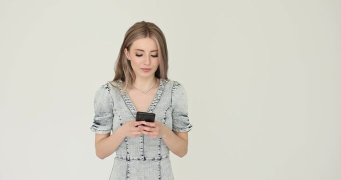 Fashionable woman carefully types message on sleek smartphone. Female in fitted denim dress writes important message against white background