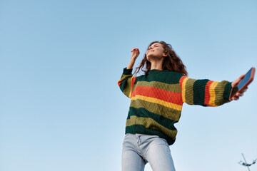 Fototapeta na wymiar Young woman in a colorful striped sweater dances with closed eyes and a smartphone in her hand against a clear blue sky. Low angle view. Concept of freedom, joy, and happy lifestyle.