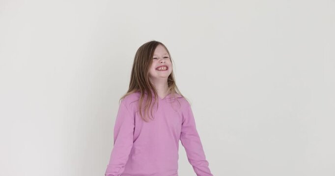 Excited little blonde girl in pink hoodie jumps up on white background. Joyful preteen child with toothy smile plays and has fun in studio slow motion