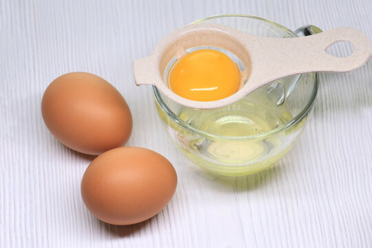 Plastic egg yolk separator with raw egg yolk sitting on glass cup with egg white and two whole eggs on light wooden table. Cooking concept.
