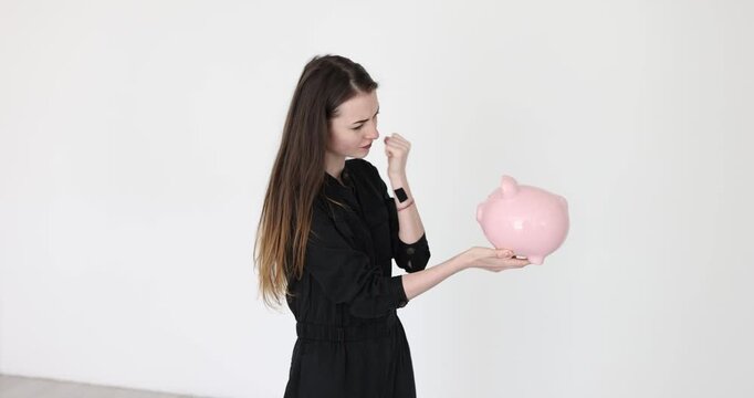 Nervous young woman threatens piggybank with index finger and fists on white background. Female suffers from money lack in studio slow motion