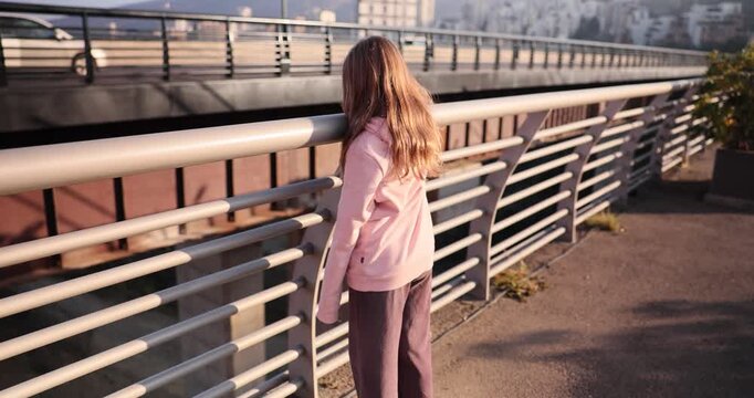 Teenage girl looks at cars driving past under city terrace backside view. Schoolgirl with loose long hair stands near footbridge railing at sunset