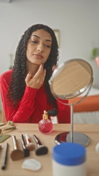 Woman touching face and applying cream at vanity mirror with brushes and perfume on table in studio; self care routine serenity.