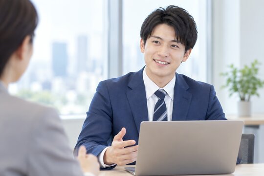 明るいオフィスでノートパソコンを前に身振りを交えて笑顔で話す日本人男性