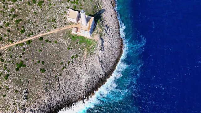 Zenithal drone zoom out revealing Santa Maria di Leuca Lighthouse on the rocky cliffs of Punta Meliso, surrounded by deep blue Adriatic waters and waves along the southern coast of Puglia, Italy.