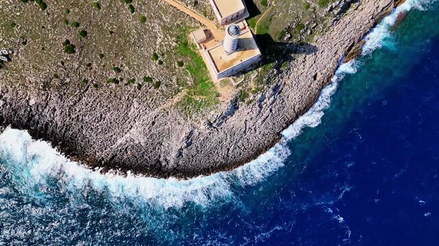 Zenithal drone spiral zoom in toward Santa Maria di Leuca Lighthouse on the rocky cliffs of Punta Meliso, with waves crashing along the Adriatic coastline in Puglia, Italy.
