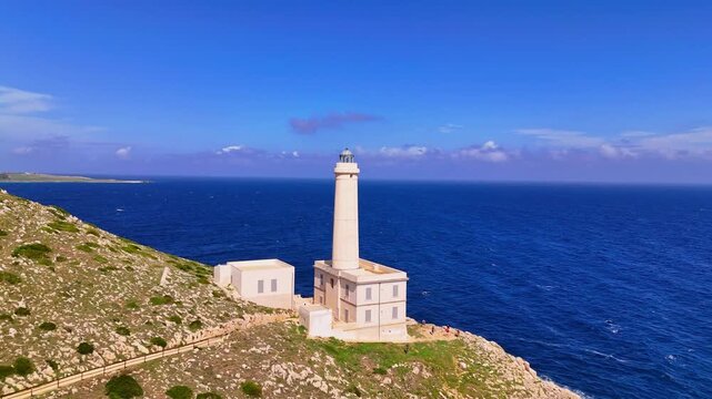 Aerial drone zoom out revealing Santa Maria di Leuca Lighthouse standing on the rocky cliffs of Punta Meliso, overlooking the deep blue Mediterranean Sea along the southern coastline of Puglia, Italy.