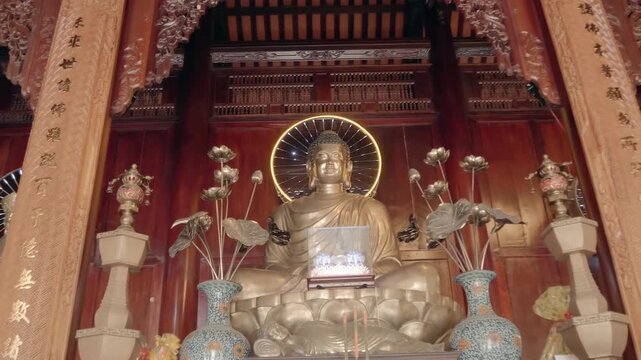 An altar with gilded bronze statue of Buddha Shakyamuni sitting in the meditation posture with vases and lotus flowers in front of the altar. Hai Duc Buddhist Institute located on Trai Thuy Hill in Nh