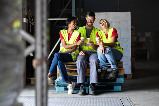 Warehouse workers enjoying a coffee break, sitting on pallets and laughing together in a factory environment