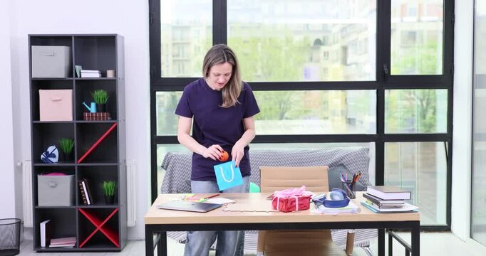 Blonde woman puts small gifts into paper bags in home office. Female in casual attire prepares presents for friends and colleagues at wooden table