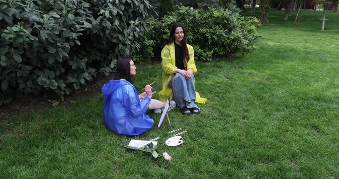 Woman draws portrait on small canvas as girlfriend sits quietly in rainy park. Females wearing plastic coats engage in art activity on green lawn