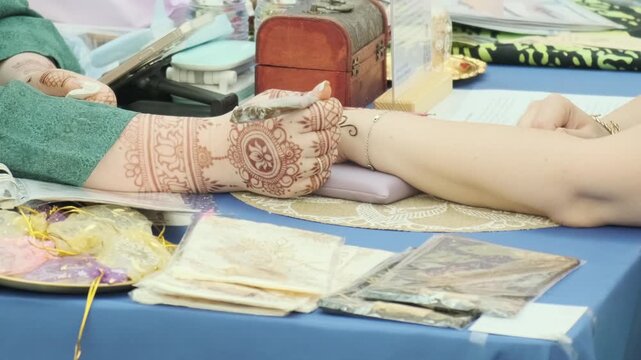 Applying traditional Indian mehndi body art with henna paste, as intricate floral and paisley pattern decorates resting forearm in bright salon setting, with ornamental bands and central floral motif