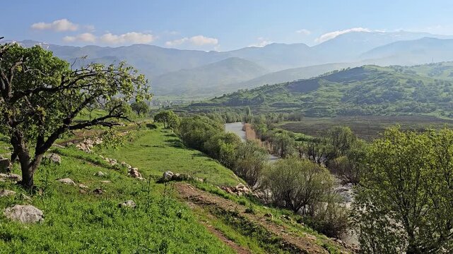 Flowing river winds through a lush green valley lined with trees and boulders, framed by distant mountains with patches of snow and clouds.
📍Ghol, Sarvabad, Kurdistan 