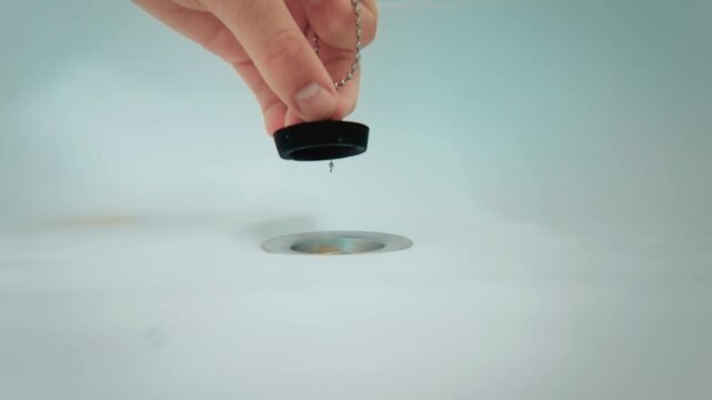 Close up hand closing bathtub drain with black rubber stopper on metal chain, view from bottom of bathtub. Fingers placing plug over drain before filling with water. Household bathroom maintenance and