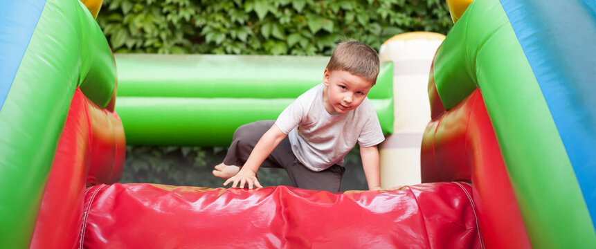 A young boy plays energetically on a vibrant inflatable slide, climbing up with excitement. The lush greenery in the background adds to the fun atmosphere.