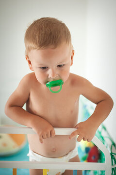 A young boy stands in his crib, wearing only a diaper and holding onto the railing with both hands. He gazes intently around the colorful nursery, filled with soft toys and cheerful decor.