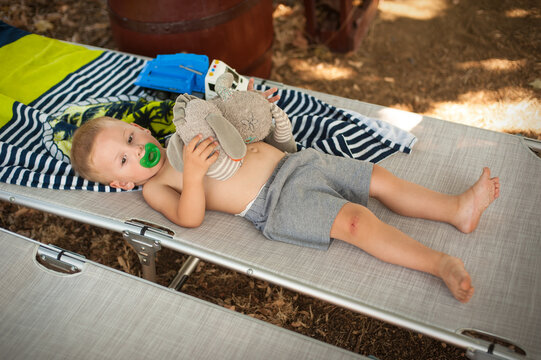 A young child lies comfortably on a cot, clutching a plush teddy bear while enjoying a peaceful moment outdoors. The warm sun filters through the trees, creating a serene atmosphere.