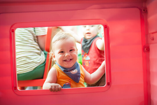A cheerful toddler peeks through a window of a vibrant playhouse, radiating joy as sunlight illuminates the scene. Nearby, other children can be seen enjoying their time.