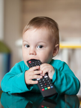 A curious toddler sits at a table, playfully biting a television remote. The room is warm and inviting, filled with soft colors and child-friendly decor, creating a playful atmosphere.