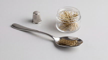 Fototapeta premium Minimalist conceptual image featuring a metal spoon filled with golden-headed sewing pins, accompanied by a thimble and a glass container of pins on a clean white surface for tailor craft.