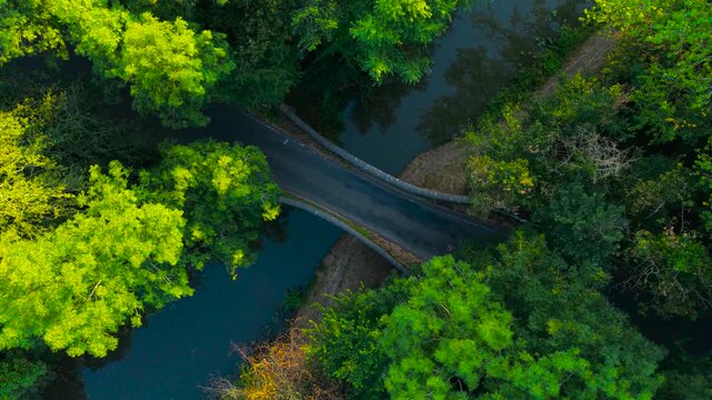 Light blue crossover driving over curved historic bridge between dense forest and tranquil river. Overhead drone perspective with colorful vehicle on remote rural road. Green woodland scenery with