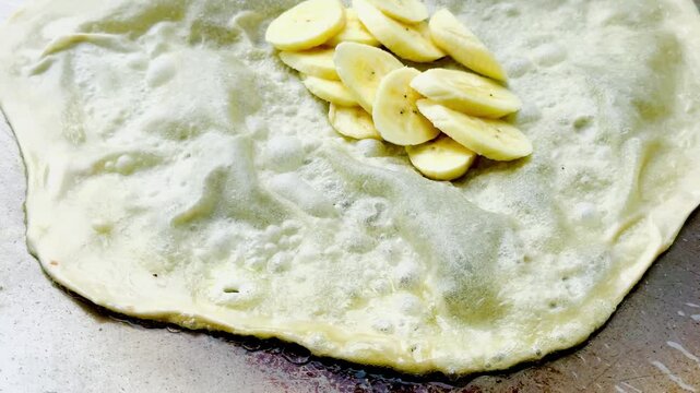Chef cooking traditional banana roti on a hot flat griddle pan. Asian street food preparation, crispy pancake dessert, sweet banana filling, and local culinary cooking process.