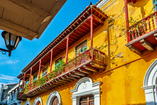 Colorful colonial architecture in Cartagena, Colombia. Bright yellow building with traditional wooden balconies adorned with blooming pink flowers against a clear blue sky.