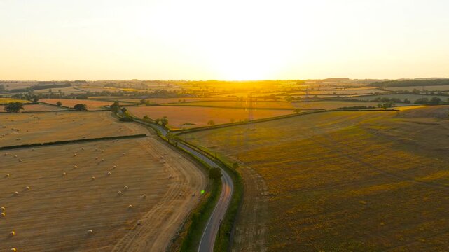 Silver crossover driving along winding country lane through harvested farmland at golden hour. Aerial view of road travel between fields and hay bales. Countryside journey across rural landscape in