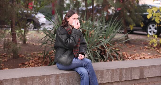Stylishly dressed woman wipes nose with disposable tissue outdoors. Female sits on wide gray parapet and cleans runny nose with tissue. Slow motion