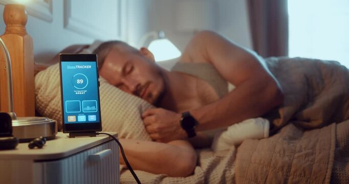 Smartphone in focus shows sleep tracking data while a young man sleeps in bed wearing a sleep tracker on his wrist in the morning.