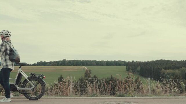 Woman walks an electric bicycle from right to left beside an open rural landscape with fields, trees and distant woodland