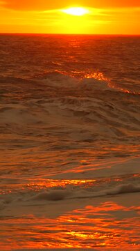 Sunset ocean waves reflecting the bright orange light of the setting sun over a serene and calm beach at golden hour