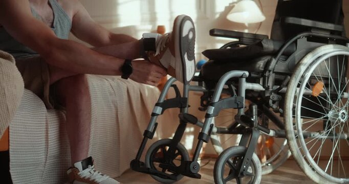 Young man with a disability sits on the bed after waking, puts on his sneakers and ties the laces beside his wheelchair as part of his independent morning routine in the bedroom.