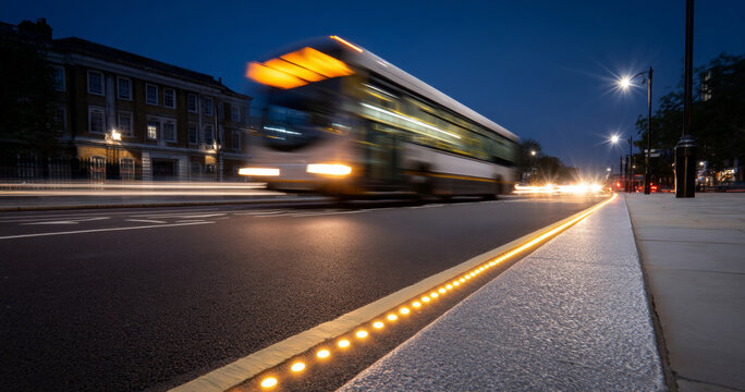 Blurred city bus speeding along illuminated urban street at dusk with historic buildings and streetlights in background
