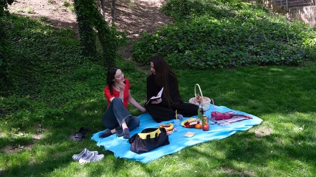 Close girl friends sit on blue blanket in park and read engaging book together. Women enjoy calm and pleasant picnic atmosphere away from noisy city