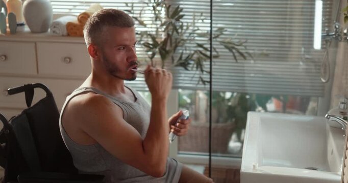 Independent young man in a wheelchair brushes his teeth in front of the bathroom mirror, completing his personal hygiene and morning routine at home.