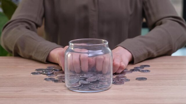 Thrifty woman drops shiny coins into transparent savings container on table. Female collects spare change for future personal goals time lapse