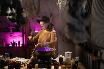 Halloween. Witch in hat with dried herbs casts spell brews a magic potion in cauldron on fire in a scary kitchen. Fantasy. © Andrii Lysenko