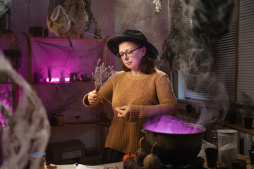 Halloween. Witch in hat with dried herbs casts spell brews a magic potion in cauldron on fire in a scary kitchen. Fantasy. © Andrii Lysenko