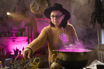 Halloween. Witch in hat with dried herbs casts spell brews a magic potion in cauldron on fire in a scary kitchen. Fantasy. © Andrii Lysenko