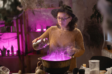 Halloween. Witch in hat adding dried herbs casts spell brews a magic potion in cauldron on fire in scary kitchen. Fantasy. © Andrii Lysenko