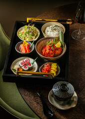 High-angle shot of a Japanese Bento lunch set served on a black wooden tray with gold handles, featuring salmon sashimi, maki sushi, crispy chicken, rice, salad, and a glass of red wine. © Simone Oppes