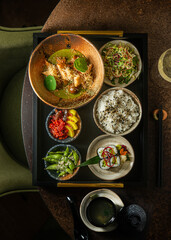 Top down view of a Japanese lunch set on a black tray featuring a green curry bowl, maki sushi rolls, rice bowl, edamame, fresh salad, side dishes, and a ceramic soup bowl. © Simone Oppes