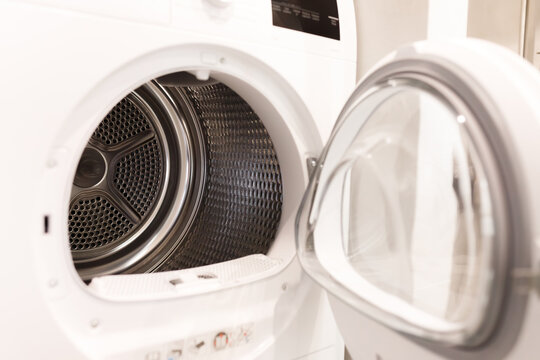 Front view of an open white modern clothes dryer machine with empty drum door swung wide background of light bathroom interior bright natural window illumination and technical household details