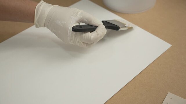 Gloved hand using a paintbrush to apply white paint or primer to a sheet of paper or board