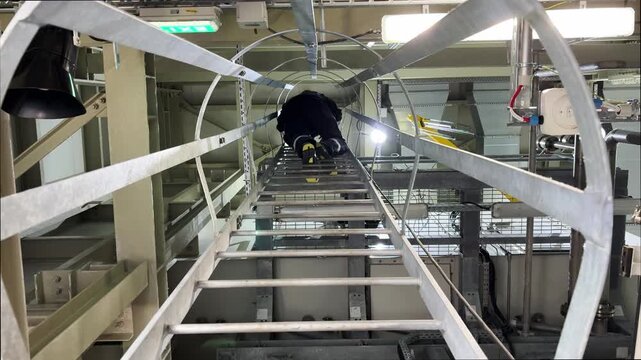 An industrial video showing a worker climbing a steep metal staircase to a high tier of an industrial facility. The footage captures the structure's towering scale, with pipes, tanks, and machinery.